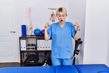 Young physiotherapist woman working at pain recovery clinic pointing down with fingers showing advertisement, surprised face and open mouth