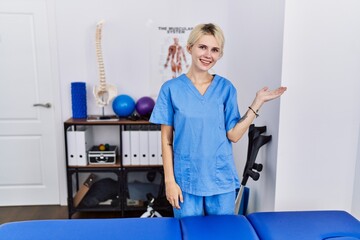 Young physiotherapist woman working at pain recovery clinic smiling cheerful presenting and pointing with palm of hand looking at the camera.