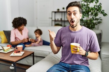 Hispanic father of interracial family drinking a cup coffee surprised pointing with hand finger to the side, open mouth amazed expression.