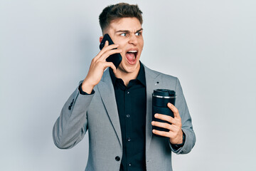 Young caucasian boy with ears dilation using smartphone and drinking a cup of coffee angry and mad screaming frustrated and furious, shouting with anger. rage and aggressive concept.