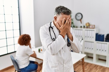 Middle age doctor man at the clinic with a patient with sad expression covering face with hands...