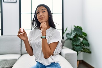 Young hispanic woman holding menstrual cup serious face thinking about question with hand on chin, thoughtful about confusing idea