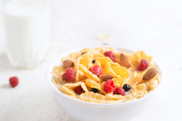 bowl of corn granola with milk, fresh raspberries, blueberries . milk in a jug and a glass  on white wooden board for healthy breakfast