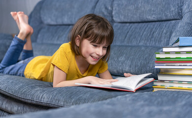 ittle girl reading a book at home. Distance learning educatiion. Cute girl liyung on sofa and read school homework