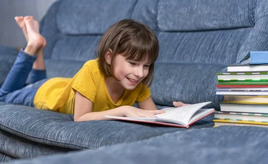 Papier Peint photo Lavable Doux monstres petite fille lisant un livre à la maison. Enseignement à distance. Jolie fille liyung sur le canapé et lire les devoirs scolaires