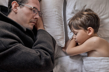 Father come to say goodbye to child boy before leave for work in morning, on bed. top view on sleepy male and kid at home, adult male in coat, in eyeglasses. family, monday, lifestyle concept