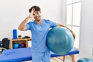 Hispanic physiotherapy man working at pain recovery clinic smiling happy doing ok sign with hand on...