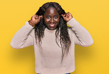 Young african woman wearing wool winter sweater smiling pulling ears with fingers, funny gesture....