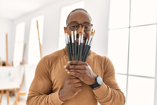 Young african american artist man covering face with paintbrushes at art studio.