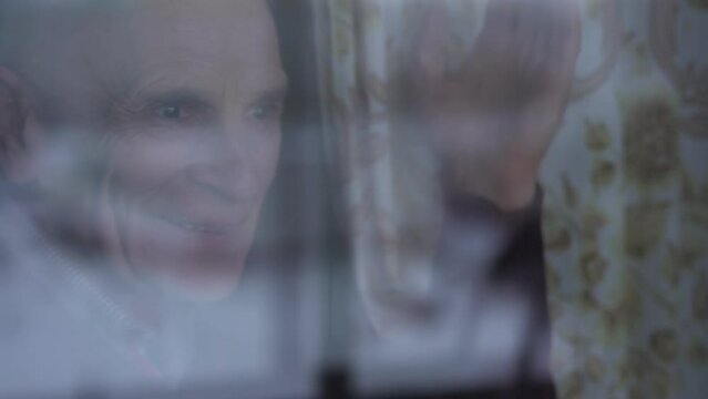 Lonely oldster standing behind window glass of hospital and waving with hand to visitors. Oldness and health care conception. Care about alone old persons.