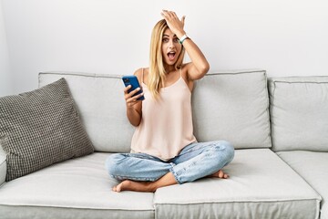 Obraz premium Blonde beautiful young woman sitting on the sofa at home using smartphone surprised with hand on head for mistake, remember error. forgot, bad memory concept.