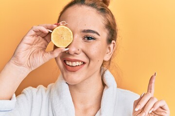 Young blonde woman wearing bathrobe holding lemon over eye smiling happy pointing with hand and finger to the side