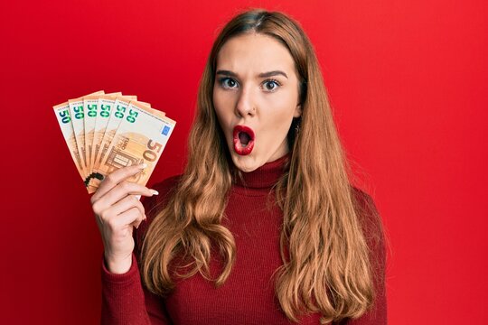 Young blonde woman holding euro banknotes scared and amazed with open mouth for surprise, disbelief face