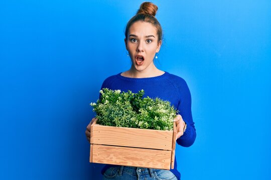 Young blonde woman holding wooden plant pot afraid and shocked with surprise and amazed expression, fear and excited face.