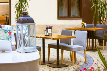 Empty cafe or restaurant terrace with wooden tables, gray chairs without people. Flowers in vases and lamps stand on tables. Green plants in pots decorate the lounge space. Place for relax outdoors.