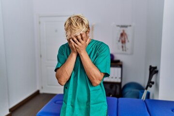 Young blond man wearing physiotherapist uniform standing at clinic with sad expression covering face with hands while crying. depression concept.
