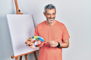Handsome middle age man with grey hair standing drawing with palette by painter easel stand winking...