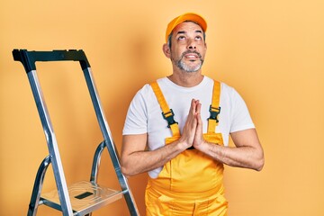 Handsome middle age man with grey hair holding ladder begging and praying with hands together with...