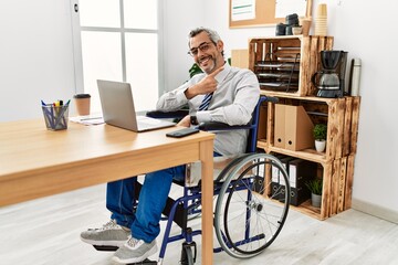 Middle age hispanic man working at the office sitting on wheelchair cheerful with a smile of face pointing with hand and finger up to the side with happy and natural expression on face
