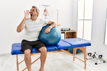 Middle age hispanic man at pain recovery clinic holding pilates ball shouting and screaming loud to...