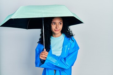 Young hispanic woman with curly hair wearing a raincoat and umbrella smiling looking to the side and staring away thinking.