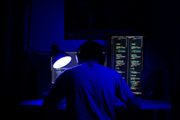 Fototapeta premium A man sits at a computer in a room at a table at night with blue lighting and programs