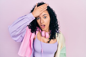 Young hispanic woman with curly hair wearing gym clothes and using headphones surprised with hand...