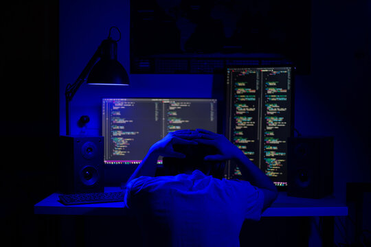 A disappointed man sits at a computer in a room at a table at night with blue lighting and programs