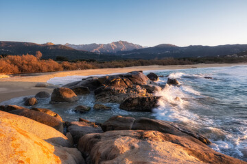 Waves crashing onto rocks at Aregno plage near Algajola in the Balagne region of Corsica with a deserted sandy beach behind and snow capped mountains in the distance