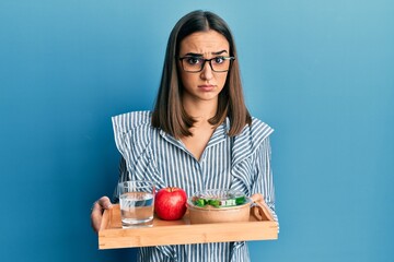 Young brunette girl holding tray with healthy lunch skeptic and nervous, frowning upset because of...