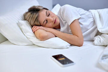 Alarm clock on smartphone lying on bed near sleepy caucasian woman to wake up on early morning, at...