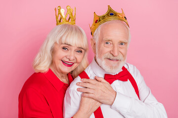 Portrait of attractive cheery gray-haired couple wearing tiara hugging isolated over pink pastel color background © deagreez