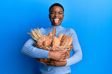 Young african american woman holding wicker basket with bread sticking tongue out happy with funny...