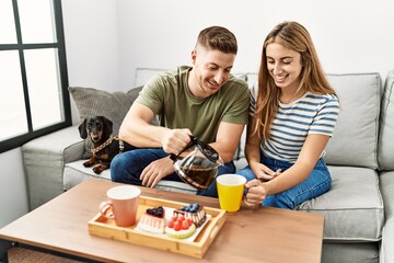 Young hispanic couple smiling happy having breakfast sitting on the sofa with dog at home.