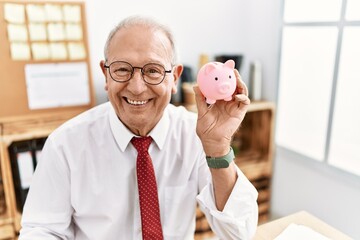 Senior business man holding piggy bank looking positive and happy standing and smiling with a...