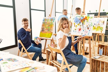 Group of middle age artist at art studio smiling with happy face looking and pointing to the side with thumb up.