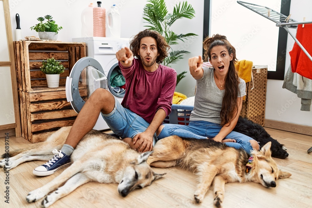 Wall mural Young hispanic couple doing laundry with dogs pointing with finger surprised ahead, open mouth amazed expression, something on the front