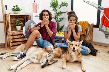 Young hispanic couple doing laundry with dogs laughing and embarrassed giggle covering mouth with...