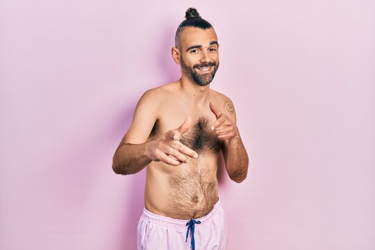 Young hispanic man shirtless wearing swimsuit pointing fingers to camera with happy and funny face. good energy and vibes.