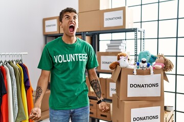 Young handsome hispanic man wearing volunteer t shirt at donations stand angry and mad screaming...