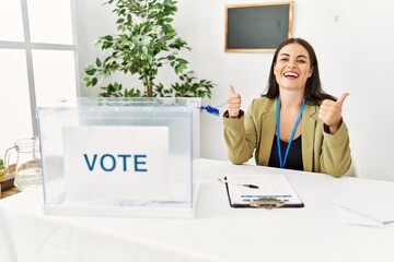 Young brunette woman sitting at election table with voting ballot success sign doing positive...
