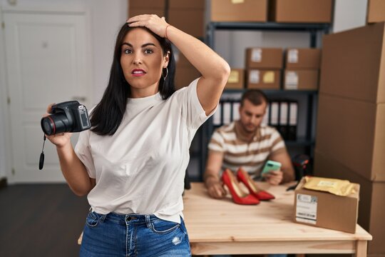 Young hispanic woman holding camera working at small business ecommerce stressed and frustrated with hand on head, surprised and angry face