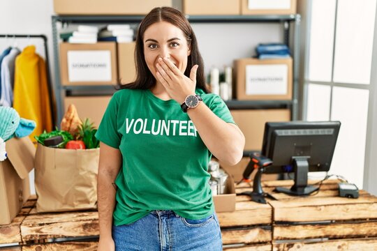 Young brunette woman wearing volunteer t shirt at donations stand laughing and embarrassed giggle covering mouth with hands, gossip and scandal concept