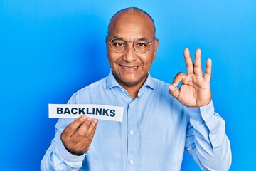 Middle age latin man holding paper with backlinks message doing ok sign with fingers, smiling friendly gesturing excellent symbol