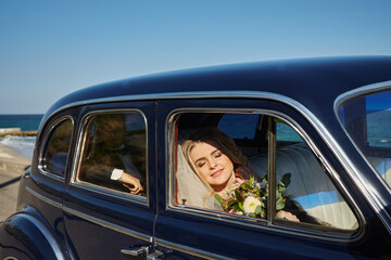 Pretty bride in luxury wedding dress sitting inside of retro car