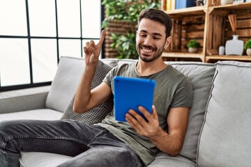 Young hispanic man using touchpad sitting on the sofa smiling with an idea or question pointing finger with happy face, number one