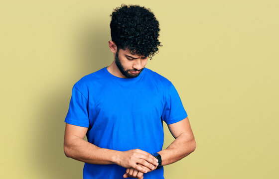 Young arab man with beard wearing casual blue t shirt checking the time on wrist watch, relaxed and confident