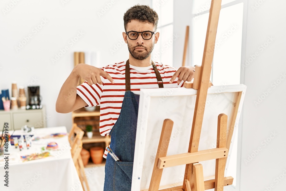 Poster Arab young man at art studio pointing down looking sad and upset, indicating direction with fingers, unhappy and depressed.