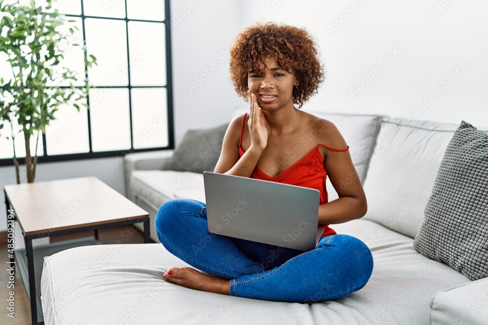 Sticker Young african american woman sitting on the sofa at home using laptop touching mouth with hand with painful expression because of toothache or dental illness on teeth. dentist