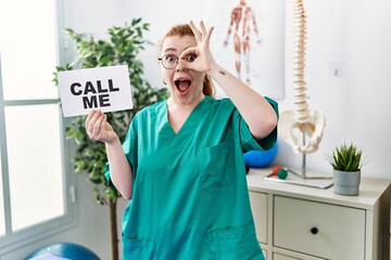 Young redhead doctor woman working at pain recovery clinic holding call me banner smiling happy...